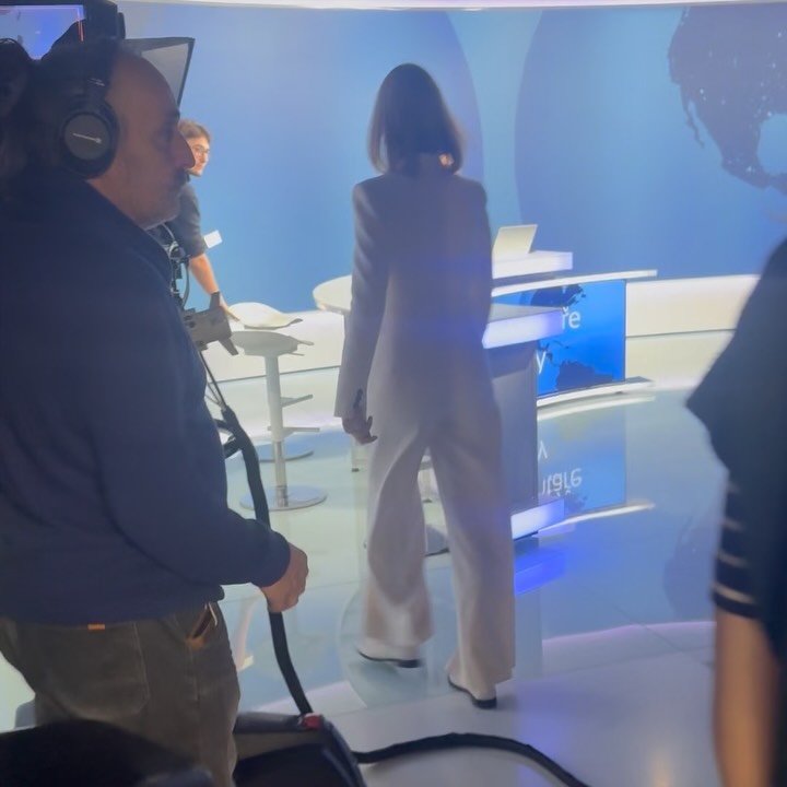 Behind-the-scenes: woman in light suit walks to news desk, crew member in foreground.