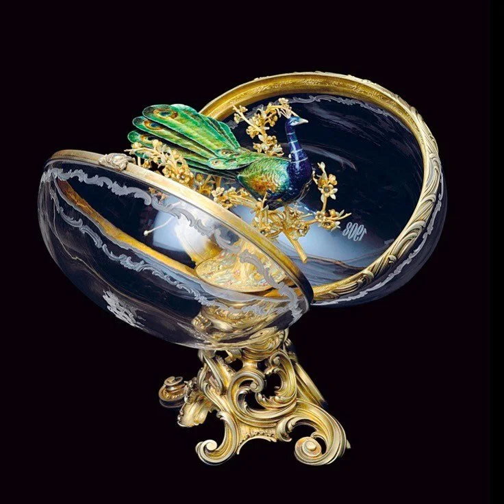 Ornate gilt and rock crystal egg, revealing a vibrant enamel peacock on golden foliage.