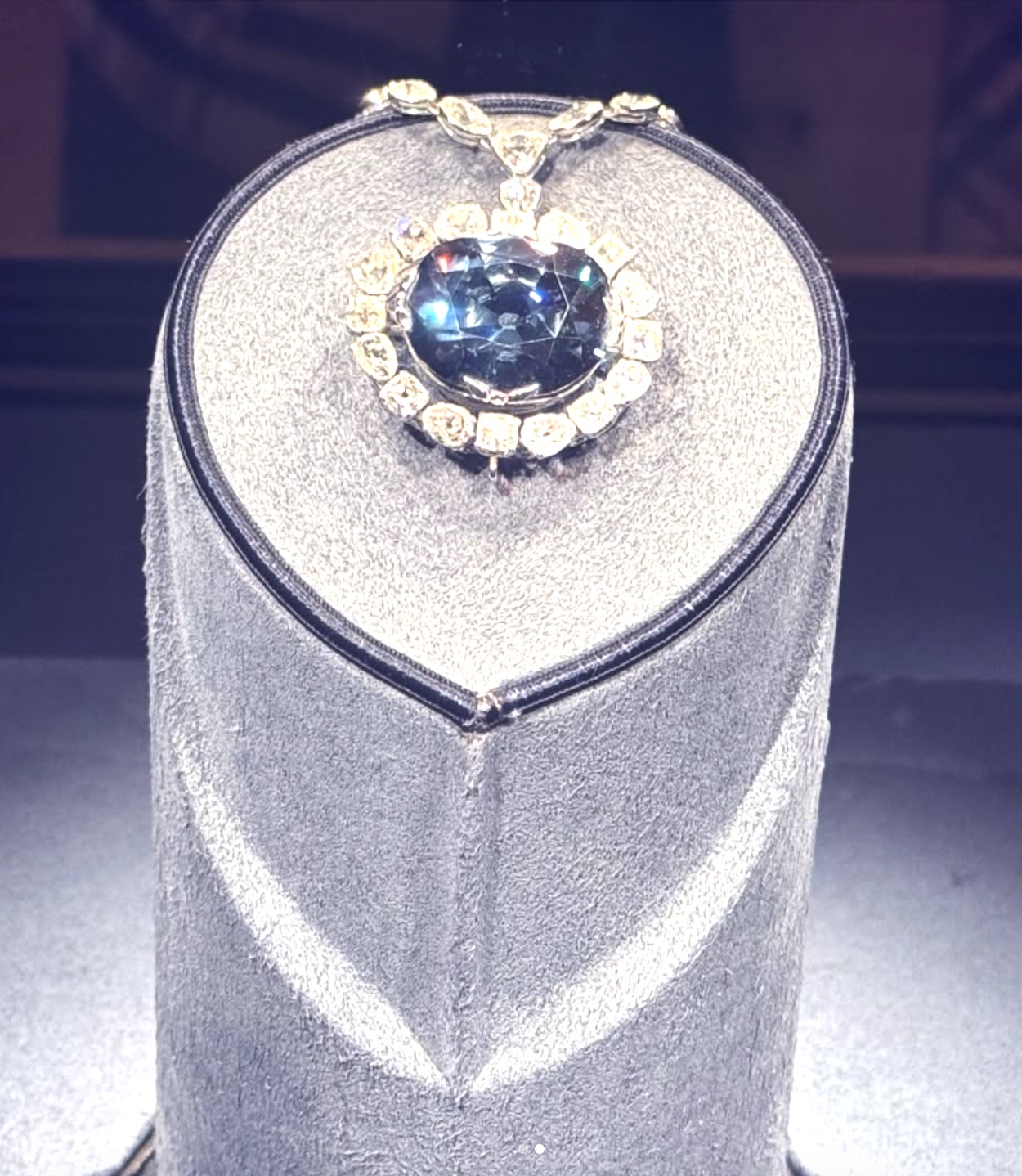 Oval dark blue diamond pendant with a halo of yellow diamonds on a grey stand.