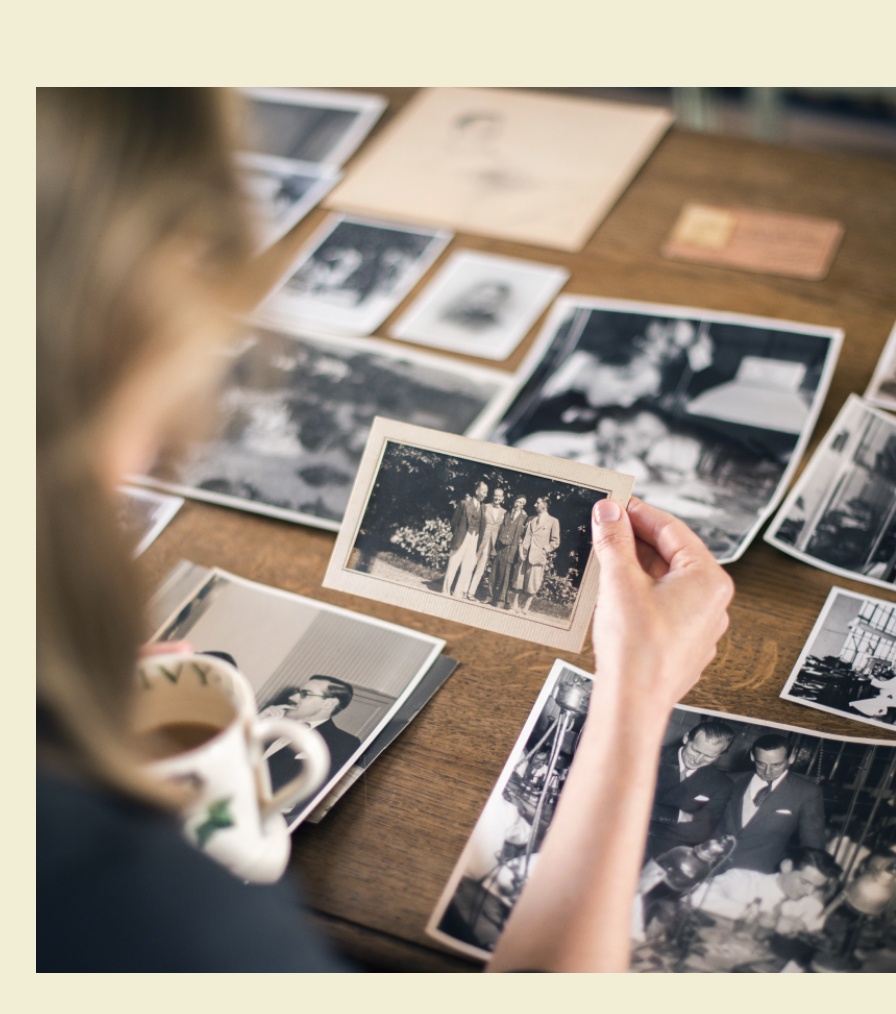 Person holding vintage black and white family photograph while sorting through collection of old photos spread on wooden table